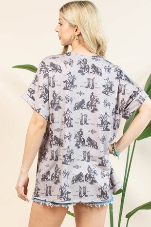 A woman shown from the back wearing a light gray oversized T-shirt dress with a black cowboy and cattle print, paired with frayed denim shorts, set against a warm beige background with green plant leaves.
