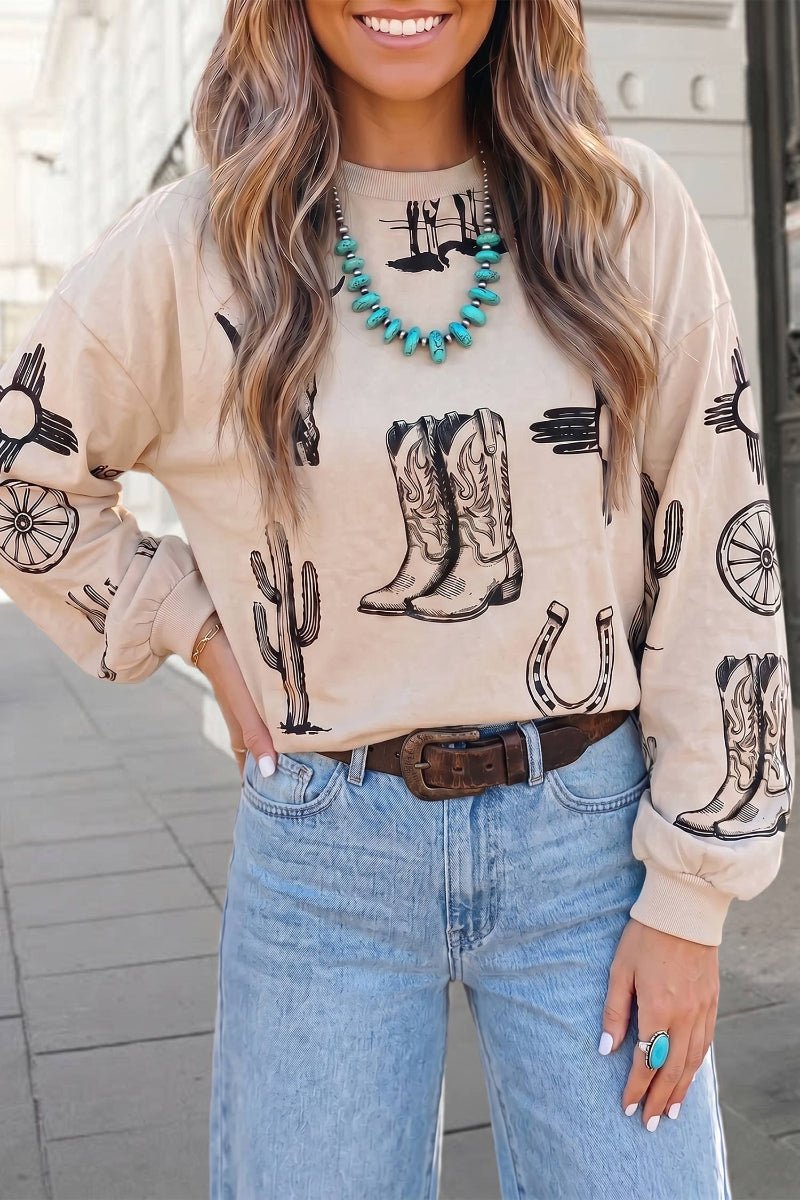 A woman wearing a beige crewneck sweatshirt with a black western print of cowboy boots, cacti, and horseshoes, paired with light blue jeans and a brown belt.