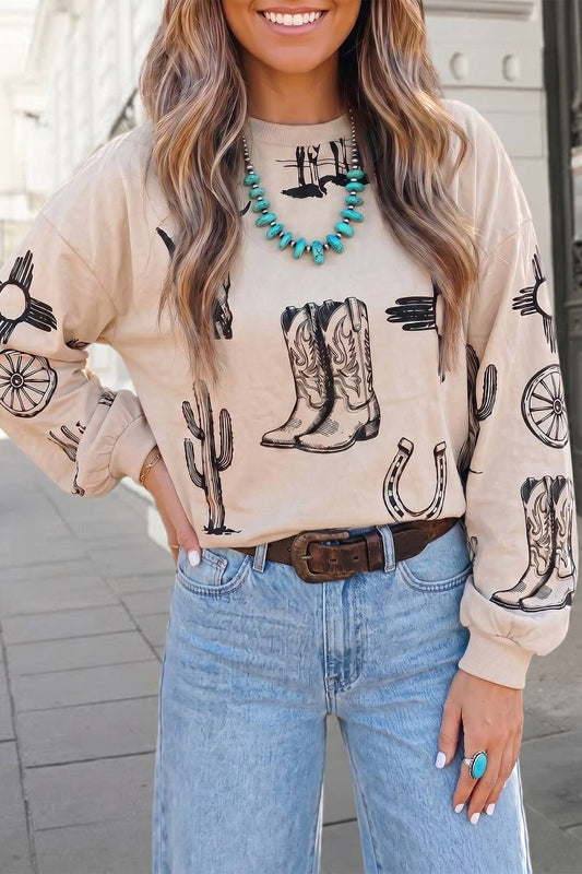A woman wearing a beige crewneck sweatshirt with a black western print of cowboy boots, cacti, and horseshoes, paired with light blue jeans and a brown belt.