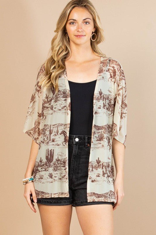 A woman modeling a lightweight open-front kimono cardigan with a desert cactus print in beige and brown, worn over a black tank top and black denim shorts against a solid beige studio background.