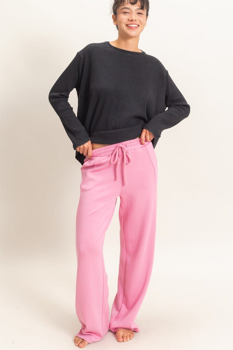 A woman modeling a black long-sleeve crewneck top and bright pink wide-leg drawstring sweatpants against a plain off-white studio background.