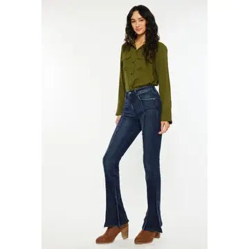 A woman modeling dark blue flared jeans with a green long-sleeve button-up shirt and brown ankle boots, standing against a plain white studio background.