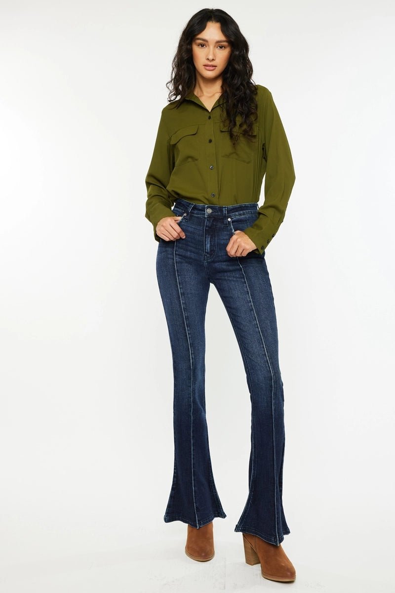 A woman modeling dark blue flared jeans with a matching olive green button-up shirt and brown boots, standing against a plain white studio background.