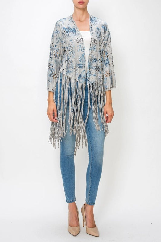 A woman modeling a light blue and gray patterned fringed cardigan over a white top and blue jeans, standing against a plain white studio background.