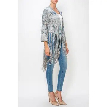 A woman models a light blue and white patterned fringed cardigan or poncho over a white top and blue skinny jeans, paired with beige heels, against a plain white studio background.