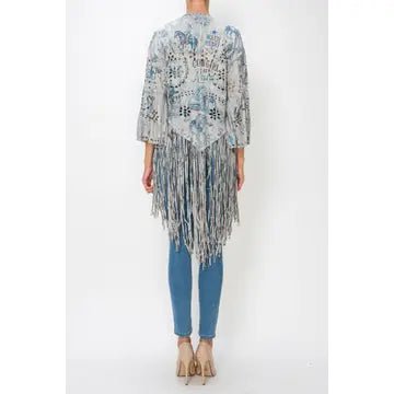 A woman shown from the back wearing a light blue and gray patterned fringed cardigan or poncho over blue jeans and beige heels, against a plain white studio background.