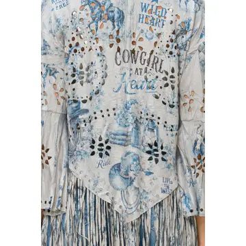 A close-up of a long-sleeve bohemian-style top featuring a light blue and white floral print, distressed details, and a tiered fringe hem. The front is decorated with text including 'COWGIRL AT HEART' and 'WILD HEART'.