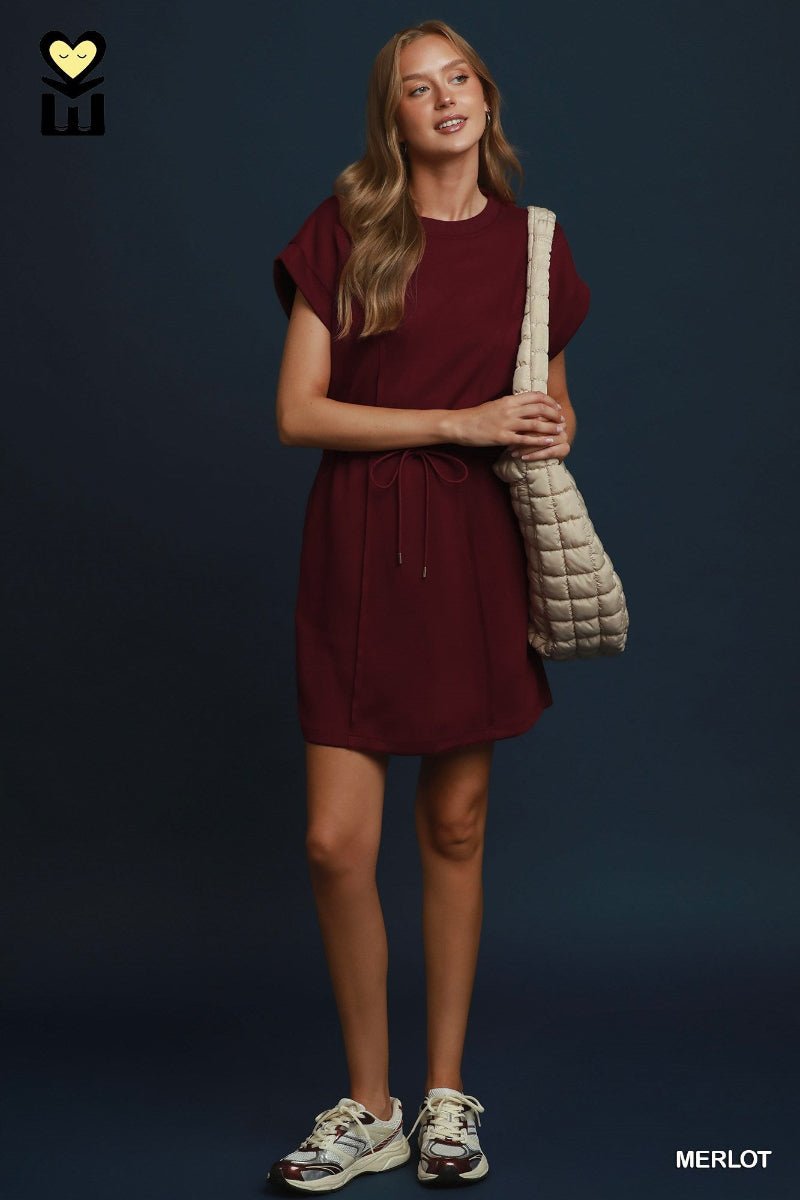A woman modeling a short-sleeve burgundy dress with a drawstring waist, carrying a beige quilted shoulder bag and wearing white sneakers, set against a dark blue studio background.