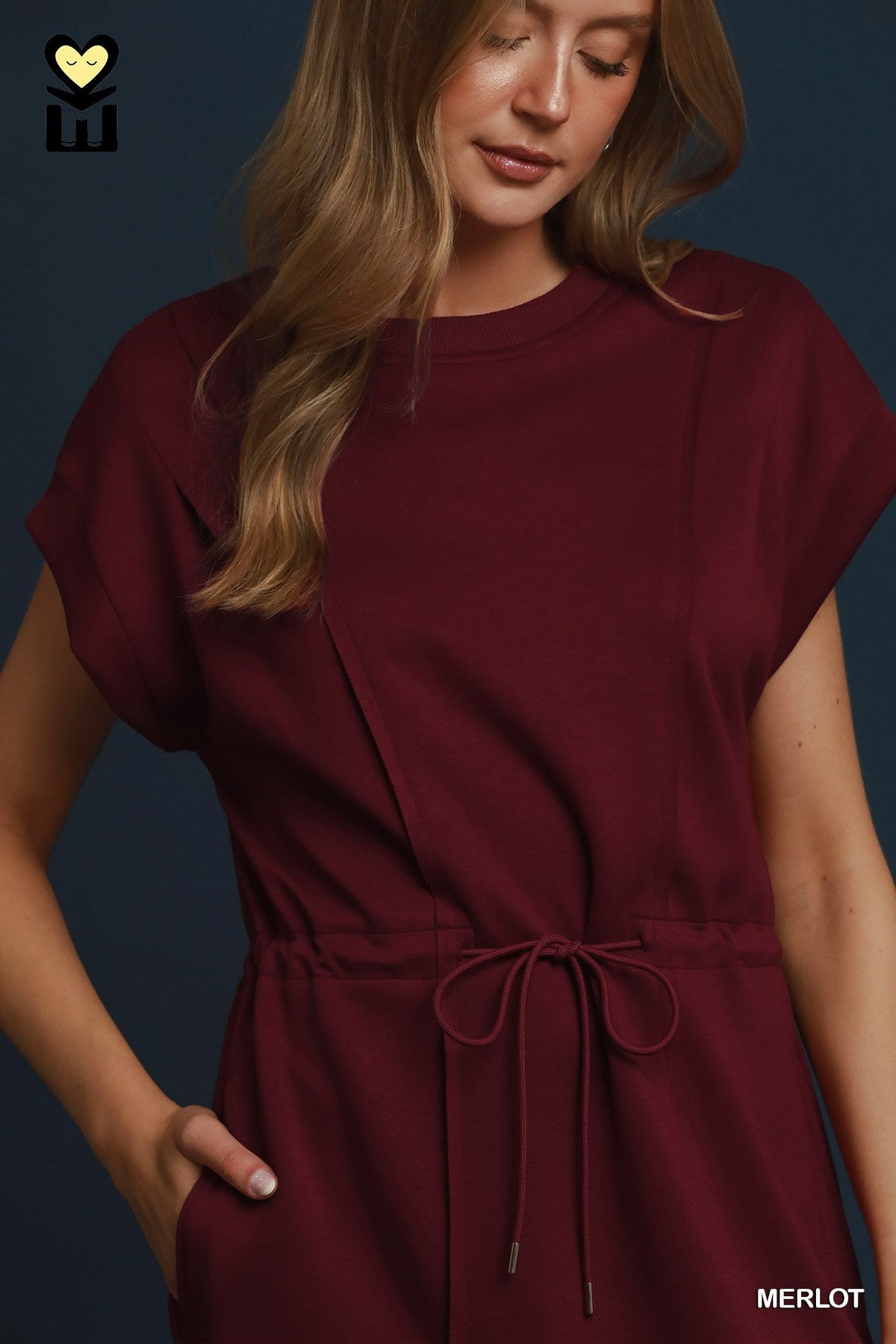 A woman wearing a short-sleeve crewneck dress in a deep merlot red, featuring a drawstring waist and side pockets, set against a dark blue background.