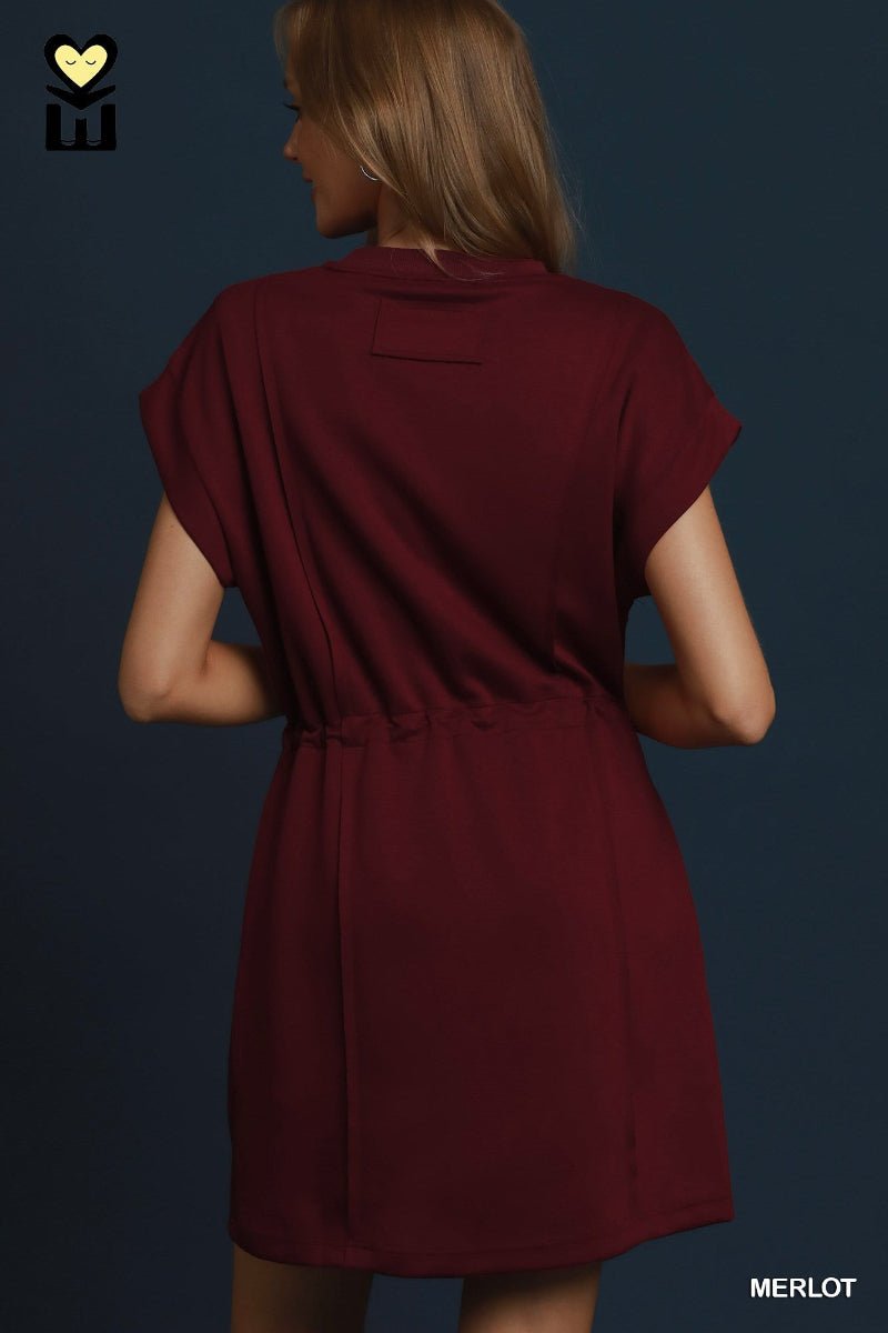 A woman shown from the back wearing a short-sleeve burgundy dress against a dark blue background. The brand logo 'EW' is visible in the top-left corner, and the color name 'MERLOT' is displayed in the bottom-right corner.
