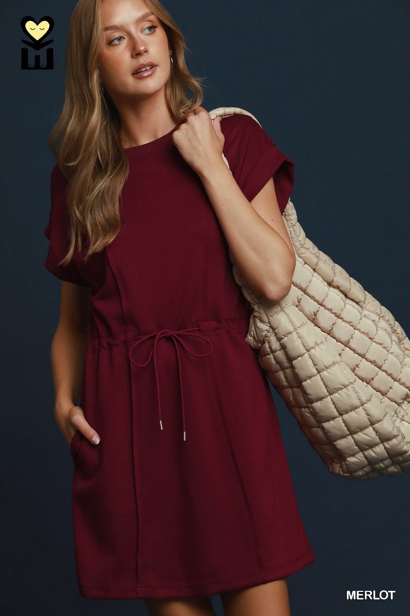 A woman models a short-sleeve burgundy dress with a drawstring waist, carrying a large quilted cream tote bag against a dark blue background.