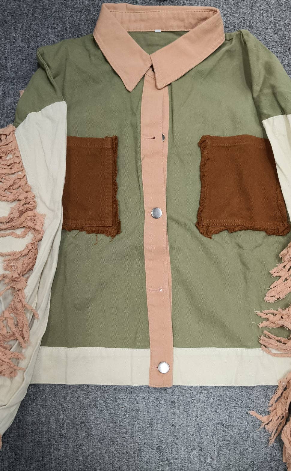 A color-blocked button-up shirt laid flat on a gray carpeted surface. The shirt features an olive green body, a tan collar and center placket, brown patch pockets, and cream-colored fringed sleeves.