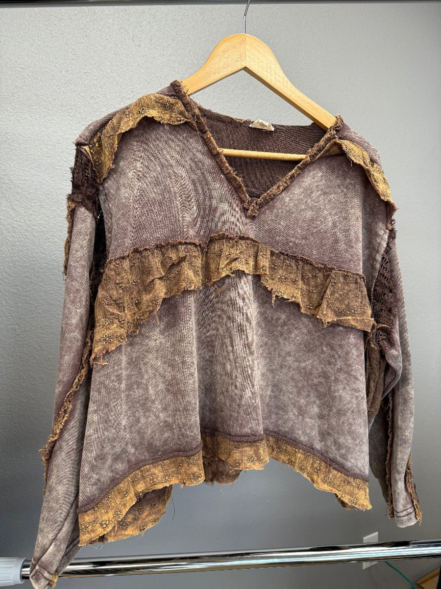 A distressed, vintage-style brown and taupe long-sleeve top with layered ruffles and frayed edges, hanging on a wooden hanger against a gray wall.