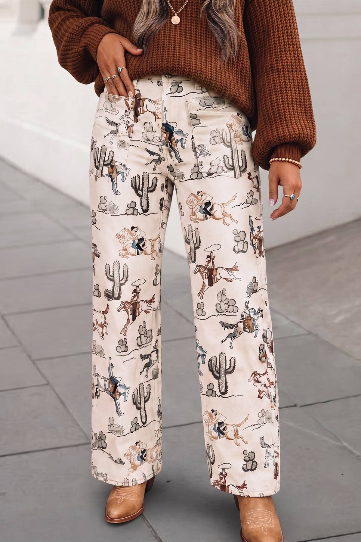 A person wearing wide-leg cream pants with a detailed western-themed print of cacti, cowboy boots, and riders, styled with a brown knit sweater and tan boots.