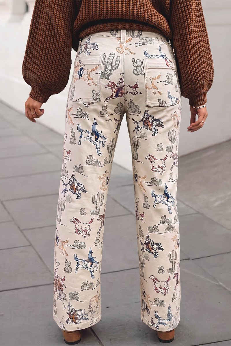 A person viewed from the back wearing cream wide-leg pants with a western print of cacti, horses, and riders, paired with a brown knit sweater and brown shoes, standing on a gray paved surface.